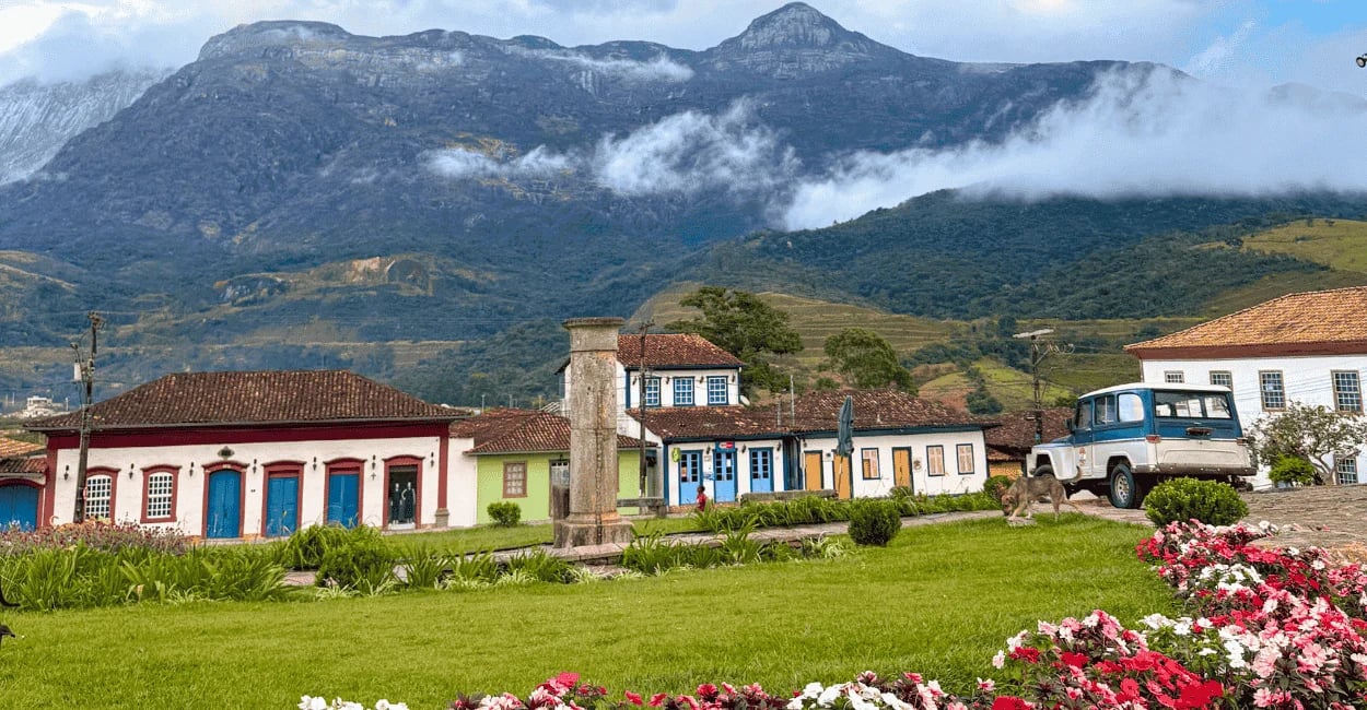 Casario colonial de Catas Altas, em MG, com praça gramada em primeiro plano e montanhas ao fundo