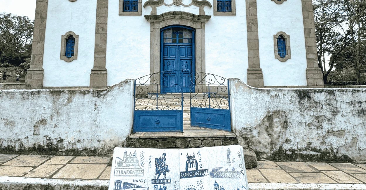 Passaporte Estrada Real em primeiro plano diante de igreja colonial de fachada branca e portas azuis