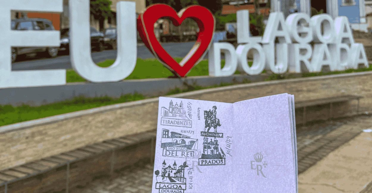 Passaporte da Estrada Real aberto, com o letreiro colorido da cidade de Lagoa Dourada ao fundo
