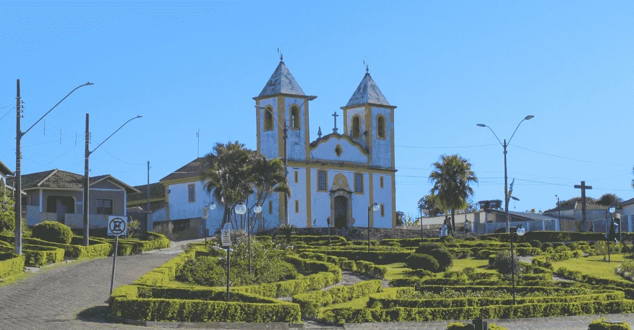 Igreja histórica em Queluzito com jardim bem cuidado, palmeiras e céu azul ao fundo