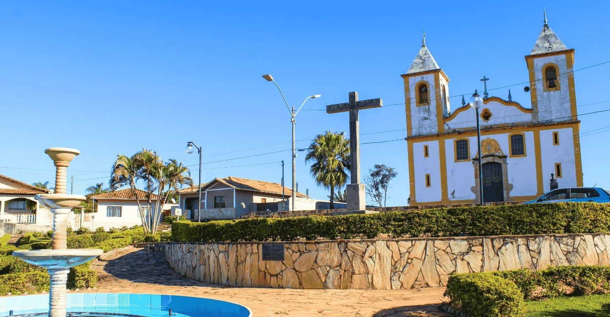 Praça com fonte e cruz de pedra em Queluzito, MG, diante da Igreja Santo Amaro sob céu azul