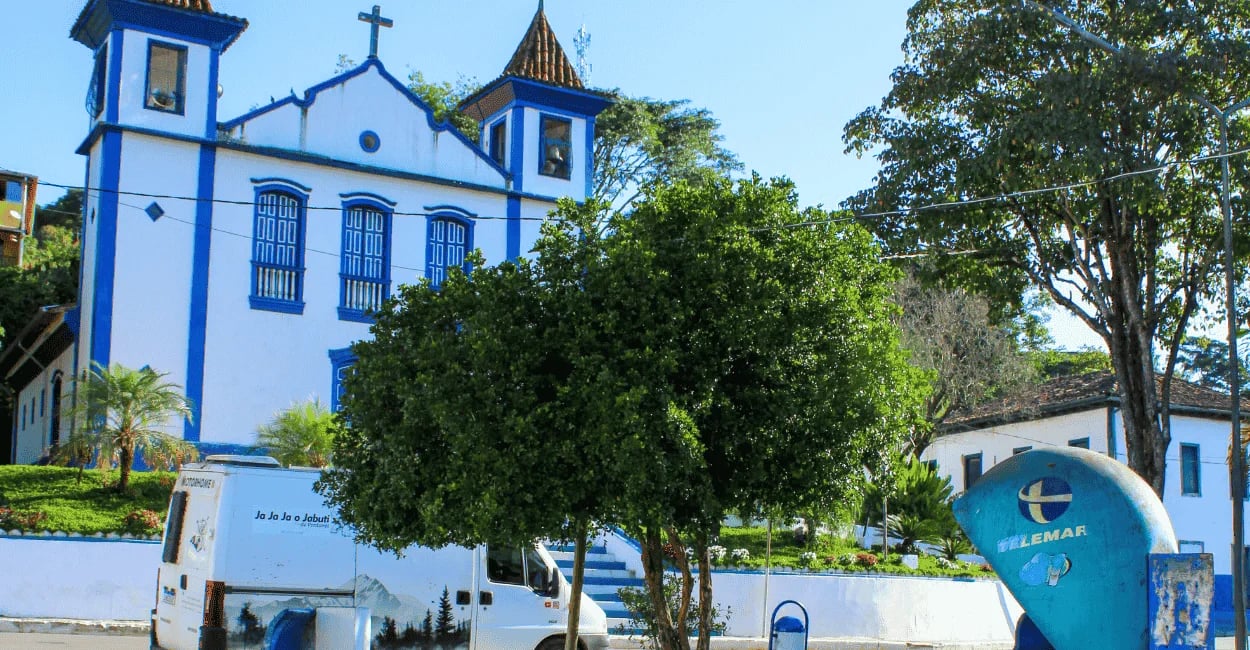 Igreja matriz branca e azul com orelhão clássico e van na praça de Ipoema, MG