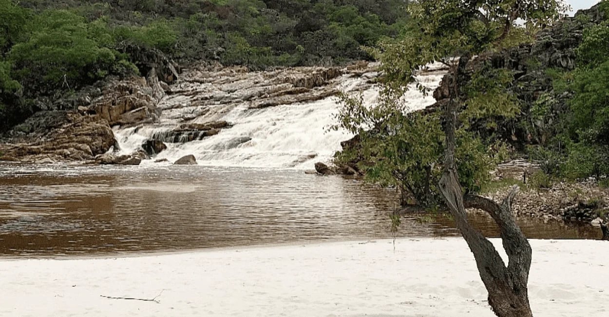 Queda d’água da Cachoeira do Telésforo com rio, areia clara e vegetação ao redor