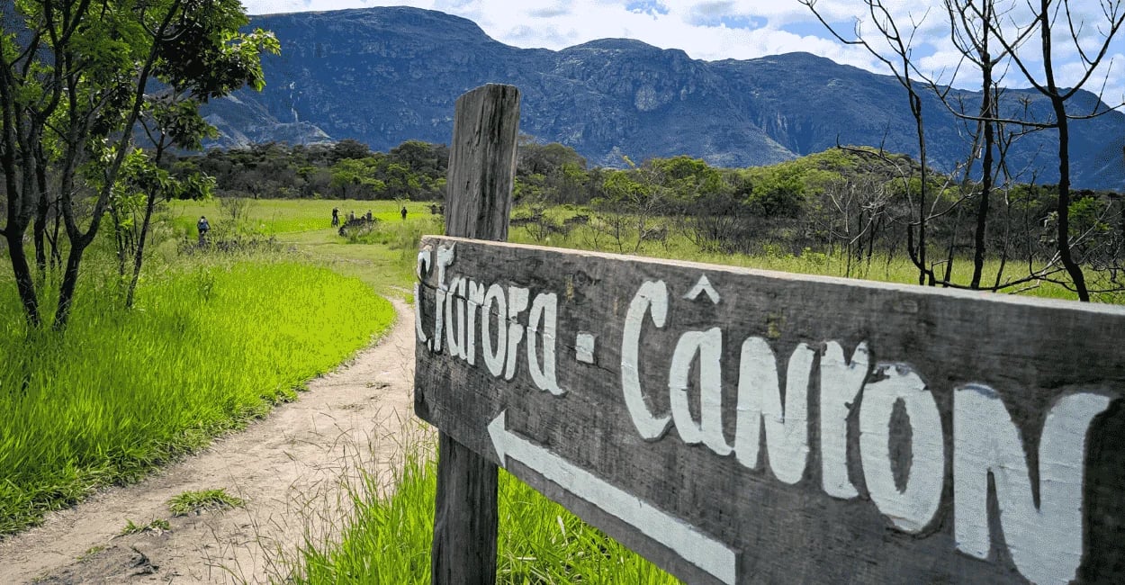 Trilha da Cachoeira da Farofa na Serra do Cipó, com placa de madeira, campo verde e montanhas ao fun