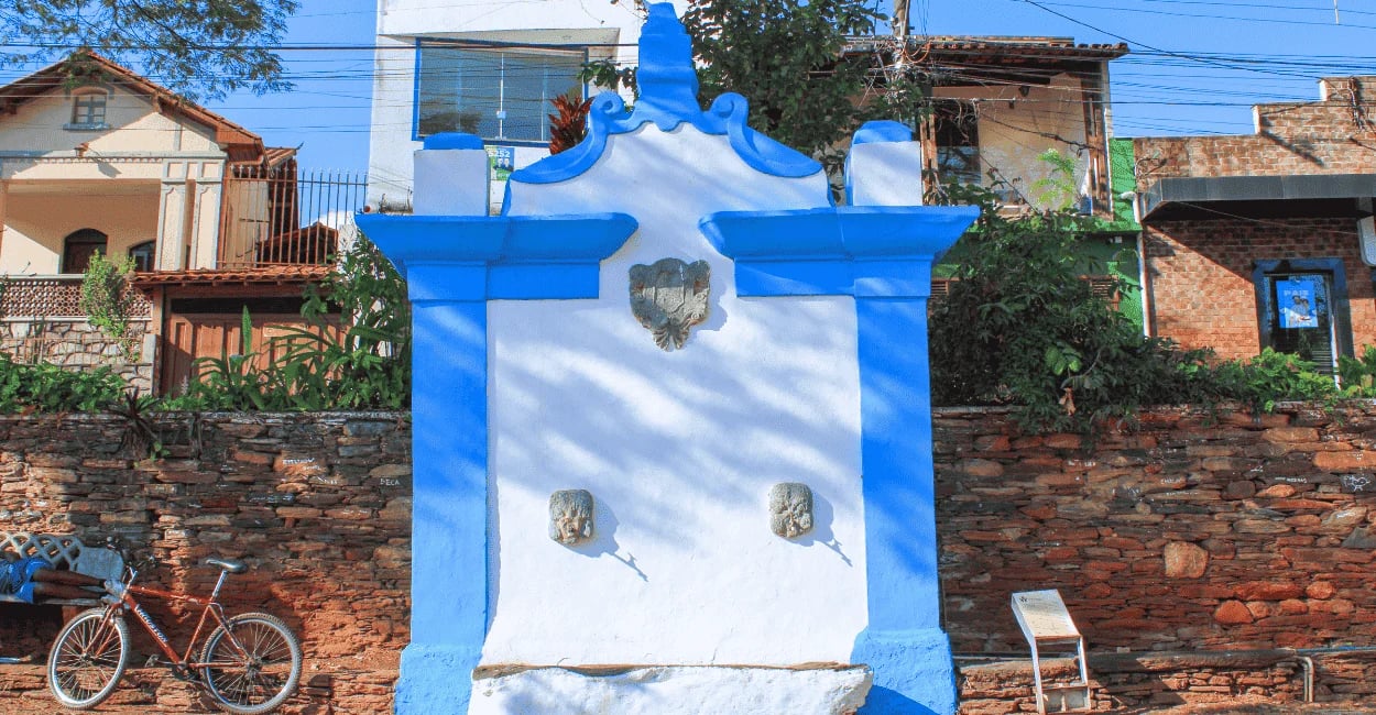 Chafariz histórico azul e branco em Sabará, Minas Gerais, diante de muro de pedra e casas ao fundo