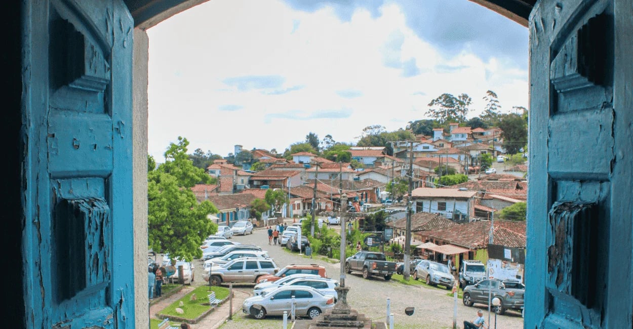 Vista da janela matriz de Lavras Novas, com casas e carros no centrino do distrito, céu nublado