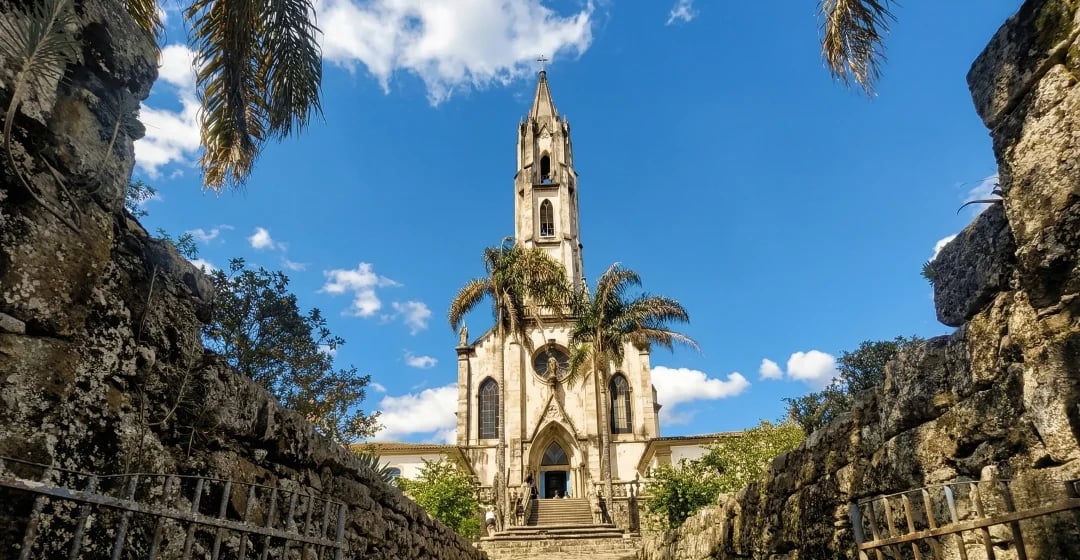 Fachada do Santuário do Caraça vista entre muros de pedra, com palmeiras, céu azul e nuvens brancas