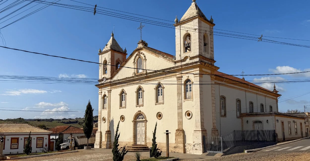 Vista lateral da Igreja Matriz de Nazareno, com duas torres, sinos e casarões históricos ao redor