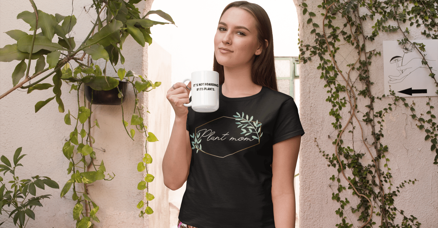 Woman holding a white plant-themed mug and wearing a black “Plant Mom” graphic t-shirt, standing near leafy green plants
