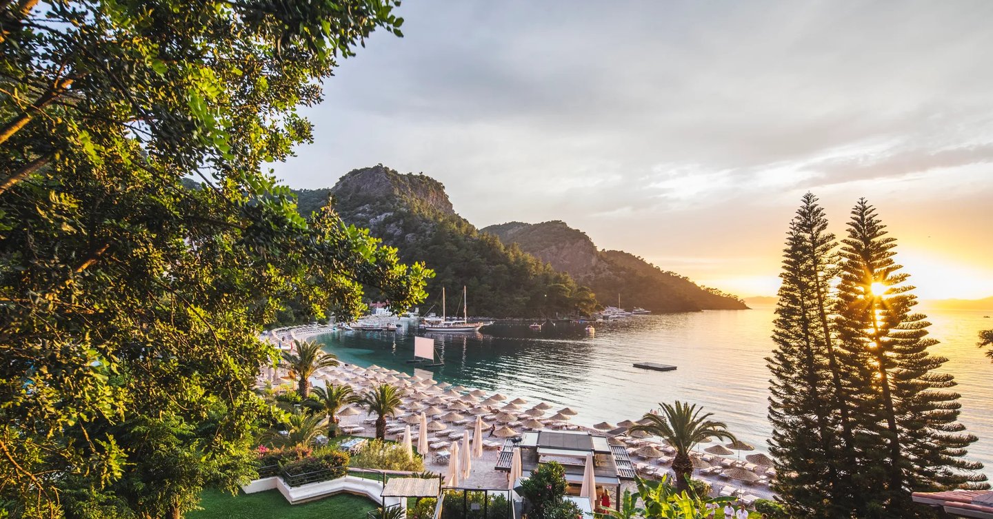Sunset view over a luxury beach resort in a tropical cove with mountains, palm trees, and ocean boats.