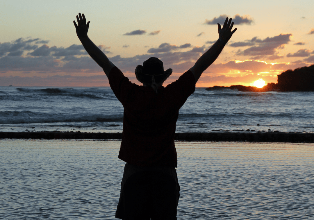 Un homme avec un chapeau qui se tiens devant un coucher du soleil a la plage 