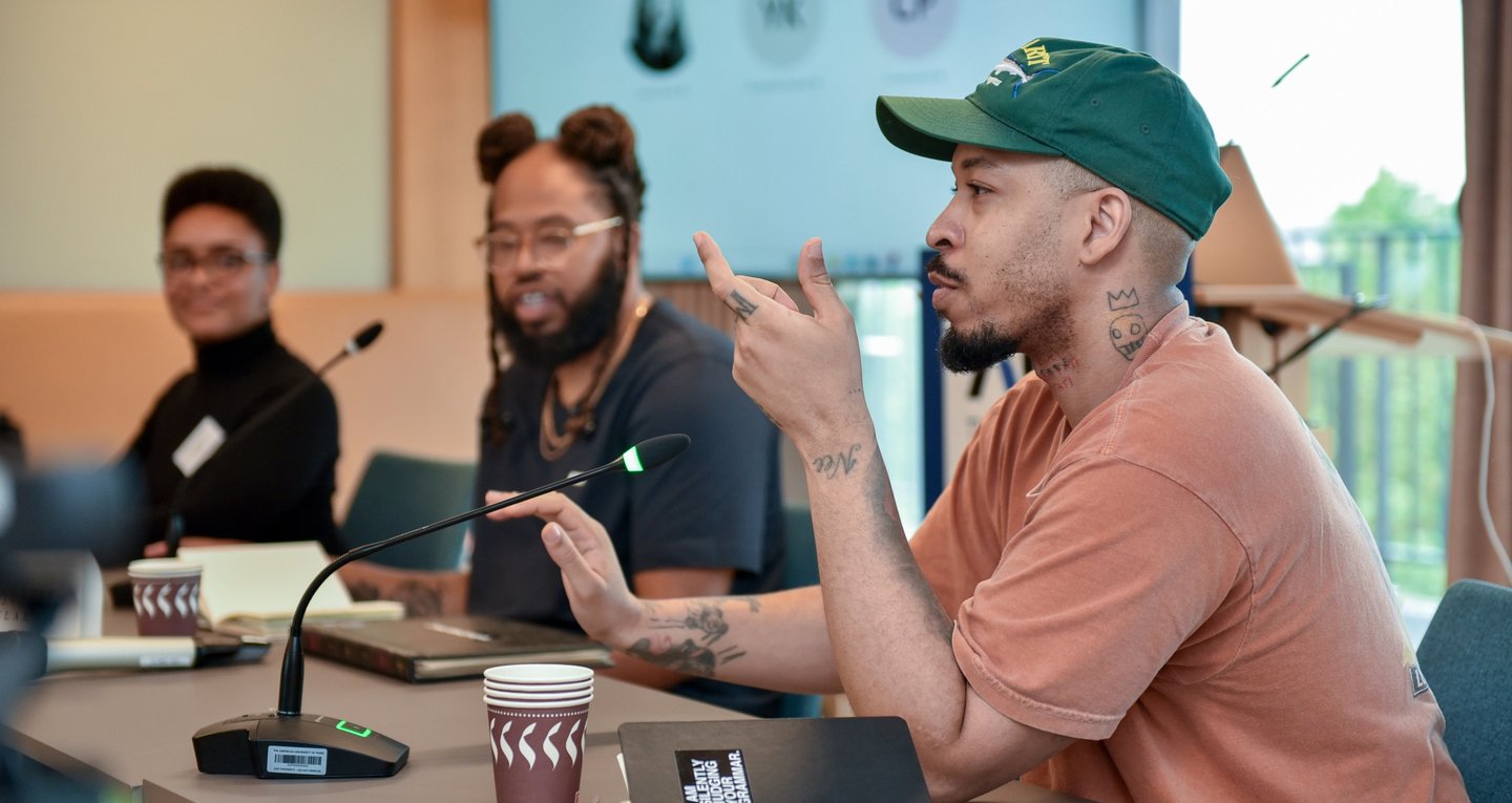 Un conférencier américain est en train de parler face à un micro, assis à côté de ses collègues.