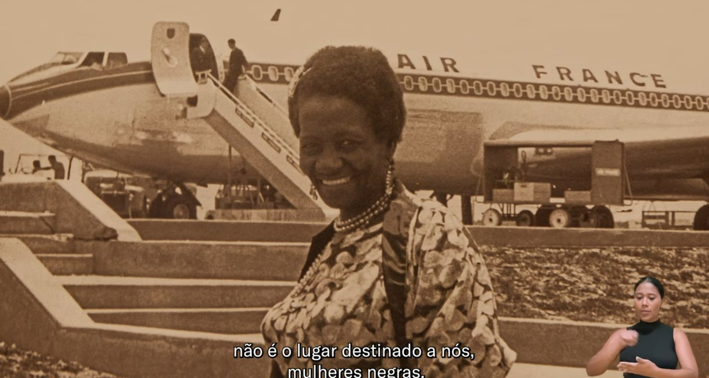Captura de vídeo da exposição de Carolina Maria de Jesus sorrindo em frente a um avião da AIR France