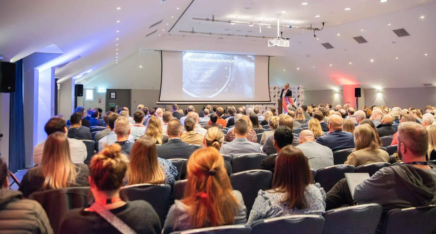 A speaker presents at a business conference with an audience seated in a large auditorium.
