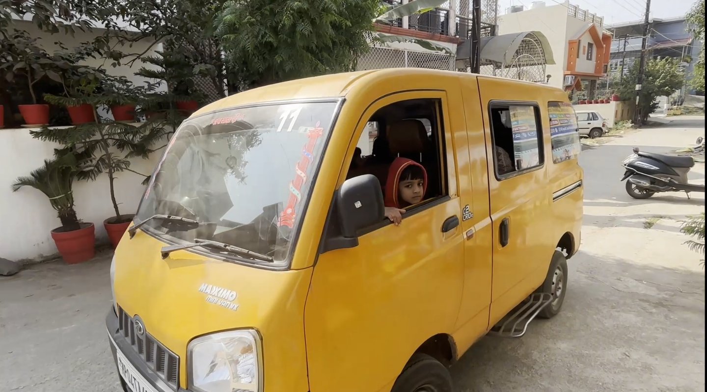 A young child in a red hoodie looking out of a yellow Mahindra Maxximo mini van on a city street.