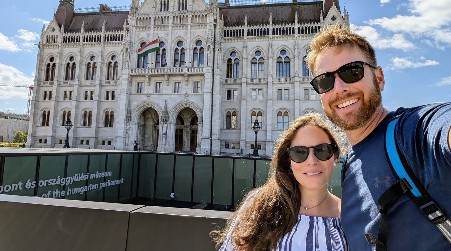A smiling couple taking a selfie in front of the neo-Gothic Hungarian Parliament Building in Budapest.