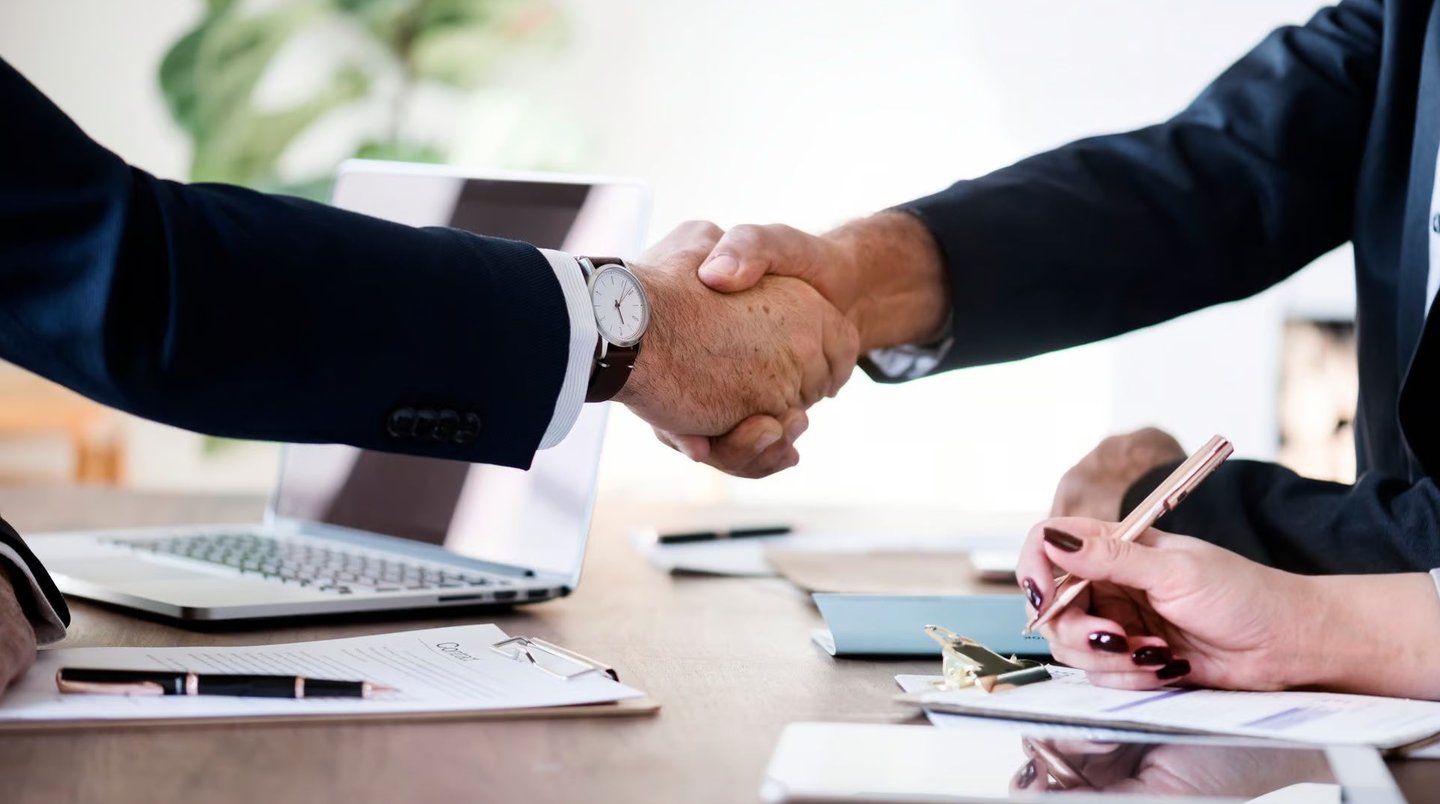 two people shaking hands over a desk with laptops