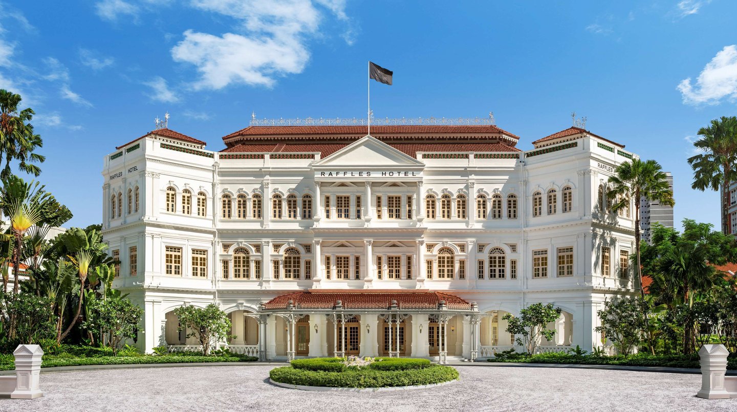 The historic white colonial facade of Raffles Hotel Singapore surrounded by tropical palm trees.