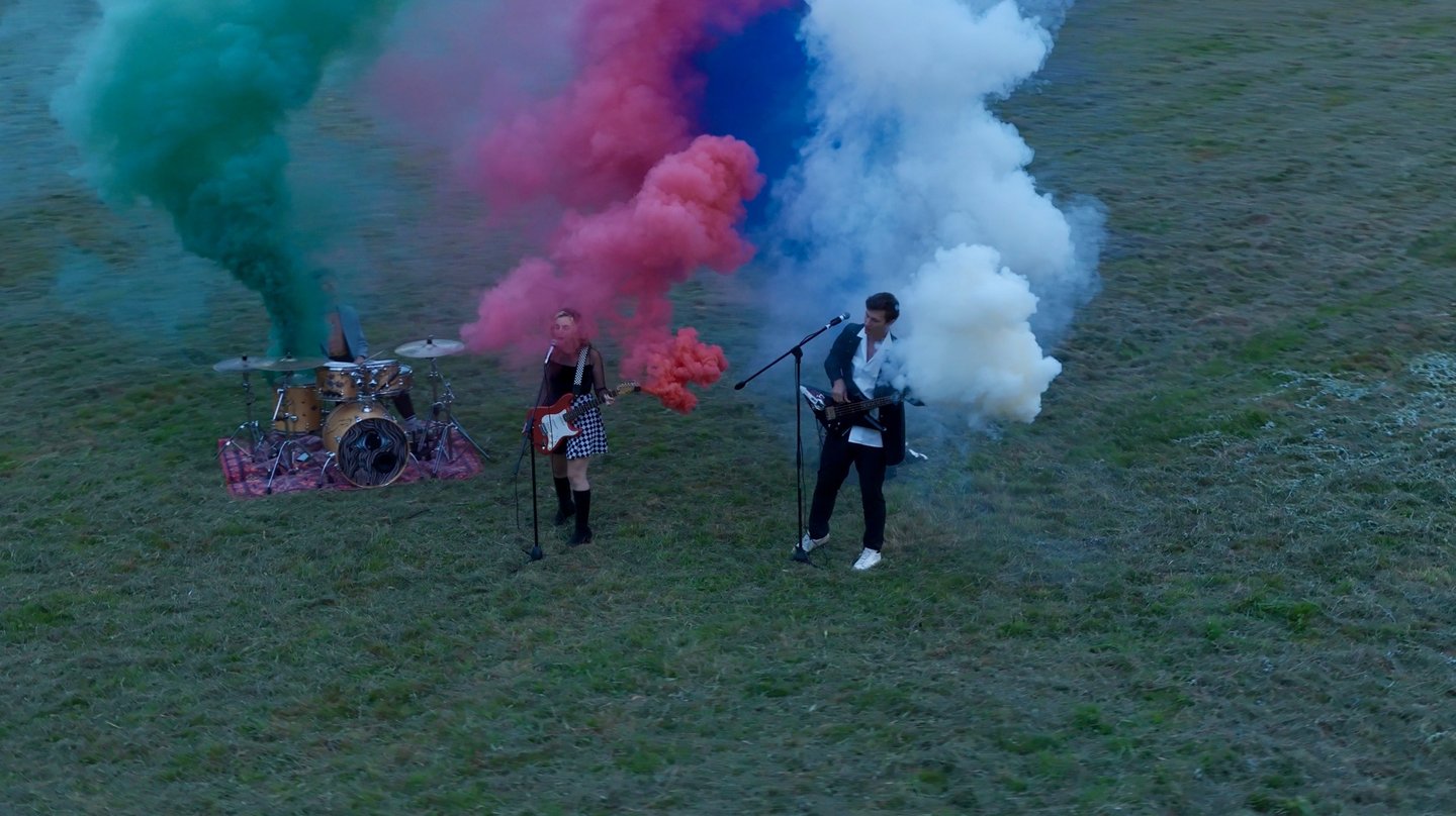 Groupe de musique de trois personnes dans un champ avec fumigènes colorés filmé par drone
