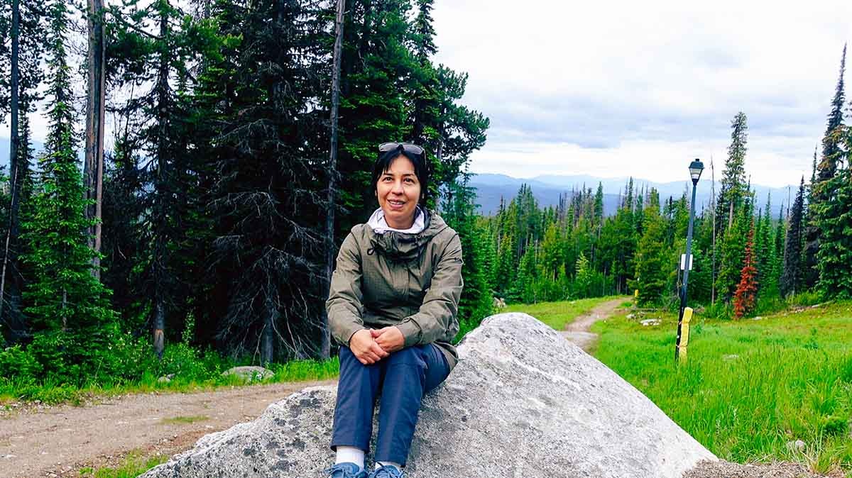 Woman sitting on a large rock beside a hiking trail surrounded by evergreen trees in Big White, Canada.
