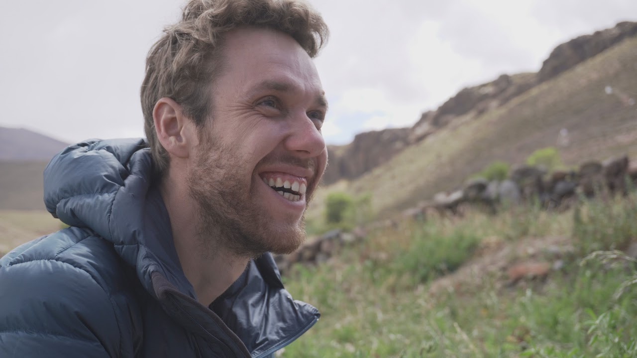 Un homme souriant, vêtu d'une doudoune bleue, en randonnée dans un paysage montagneux.