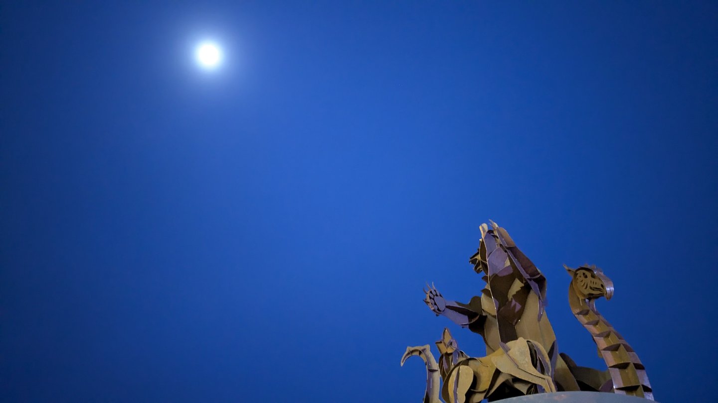 A metal bear sculpture silhouetted against a deep blue evening sky with the moon overhead.