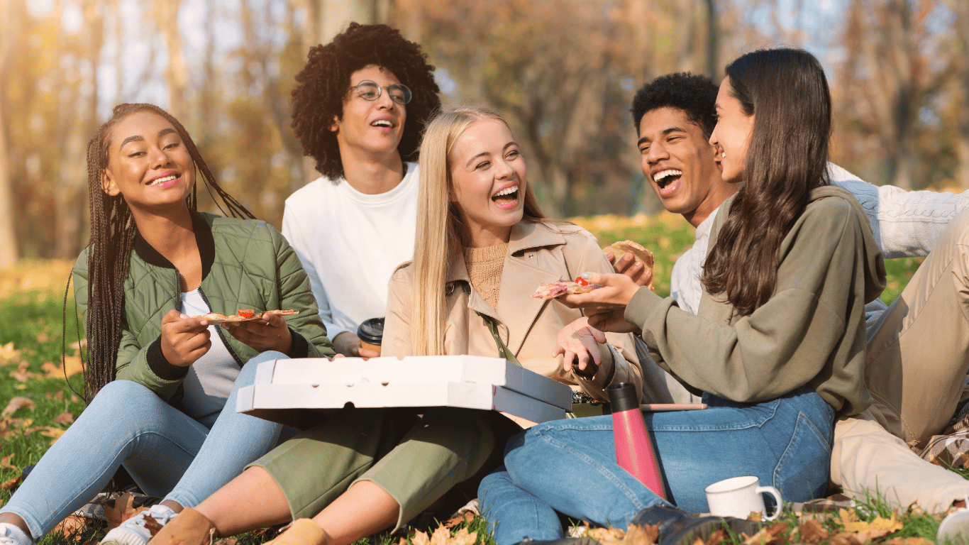 Diverse group of smiling friends enjoying a pizza picnic outdoors in a sunny autumn park.