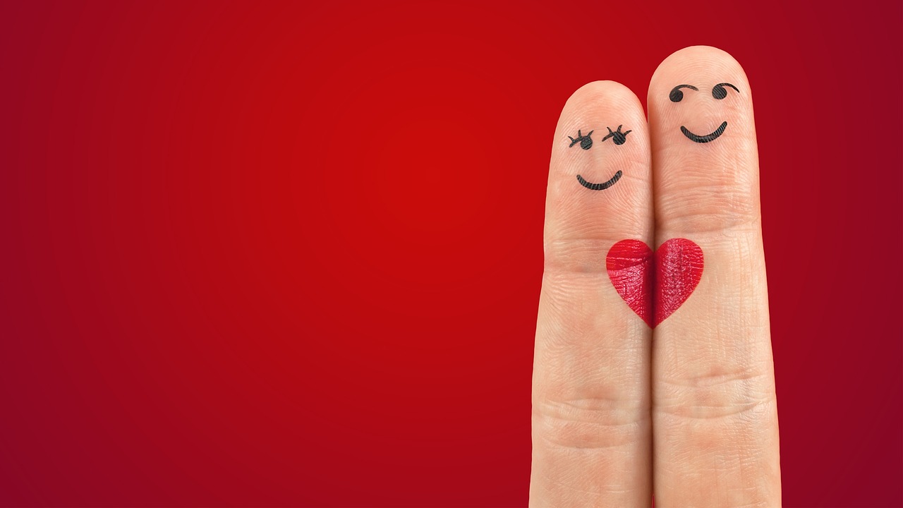 Two fingers with drawn smiley faces and a red heart represent a happy couple on a red background.