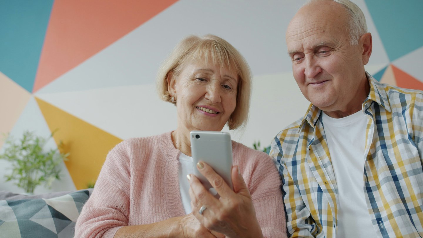 A happy senior couple sitting together on a couch while using a smartphone for a active aging