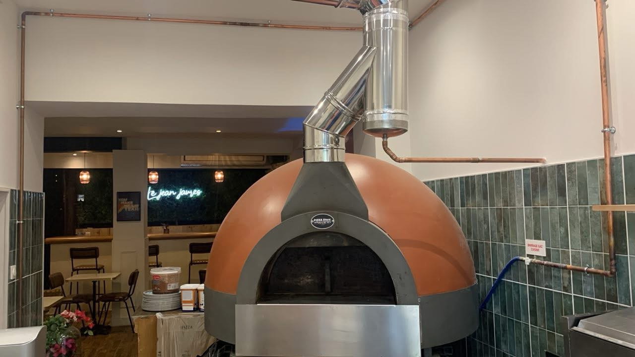 Large dome-shaped wood fired pizza oven in a modern restaurant kitchen with copper pipes.
