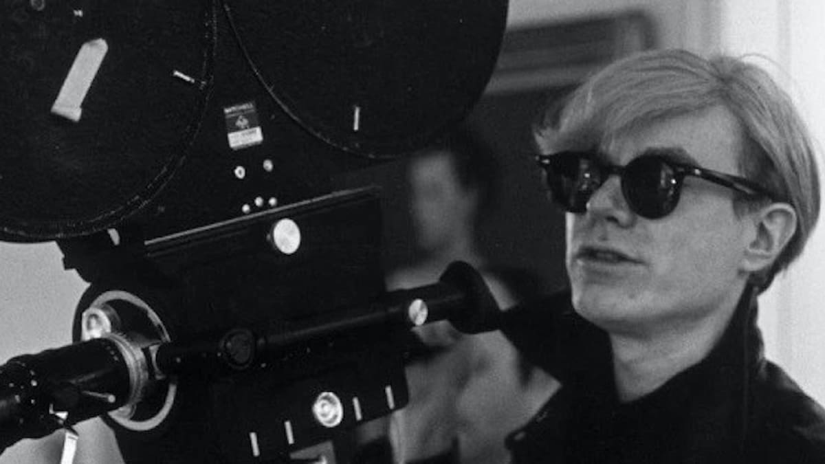 Andy Warhol segurando uma câmera de filme antiga, usando óculos escuros preta