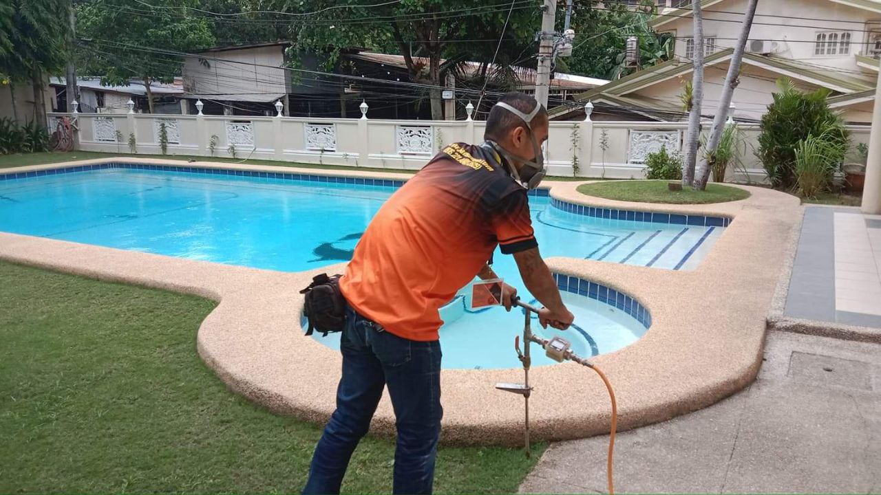 A pest control professional wearing a mask applies termite treatment near a residential swimming pool.
