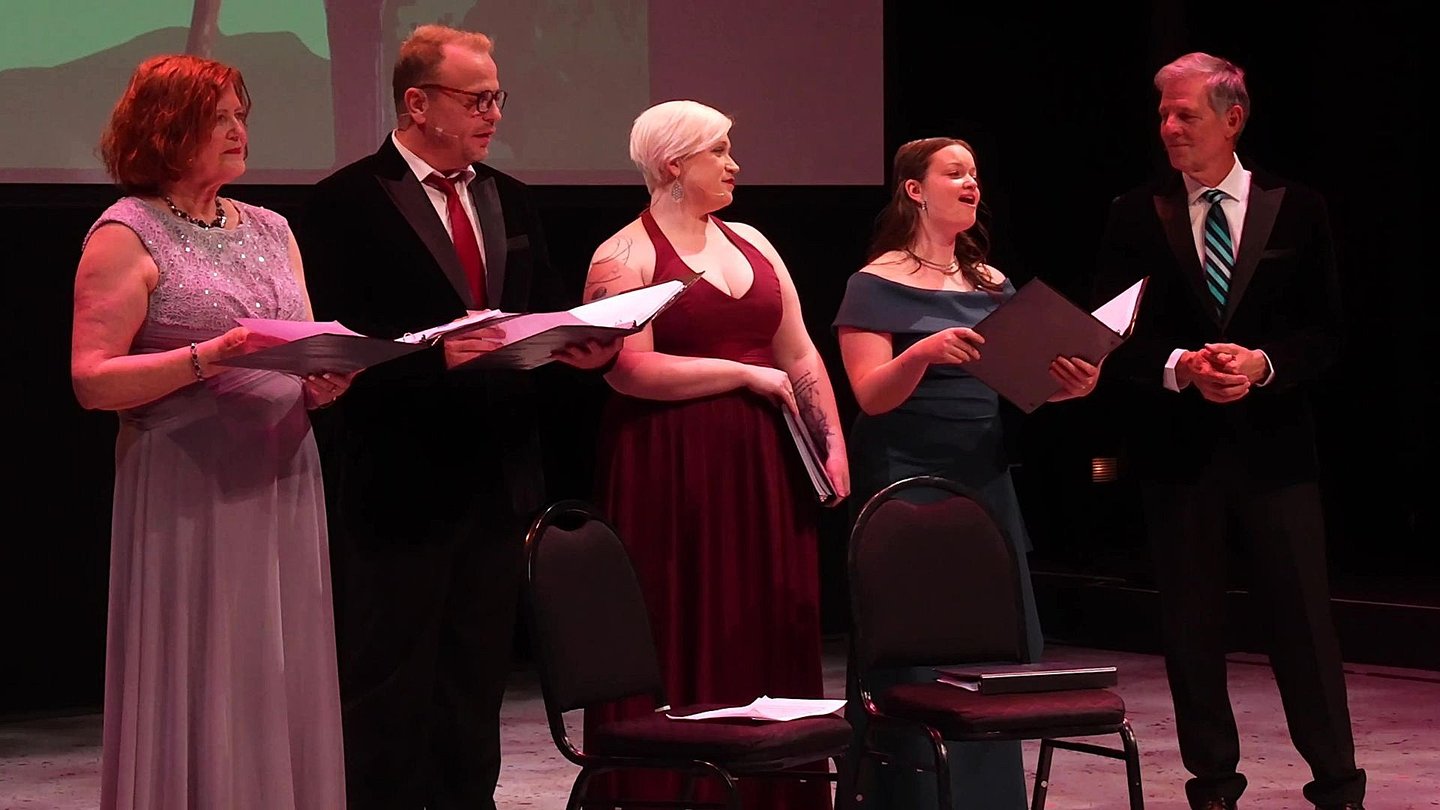 Five singers in formal attire performing on stage; four watch the soloist as she performs.