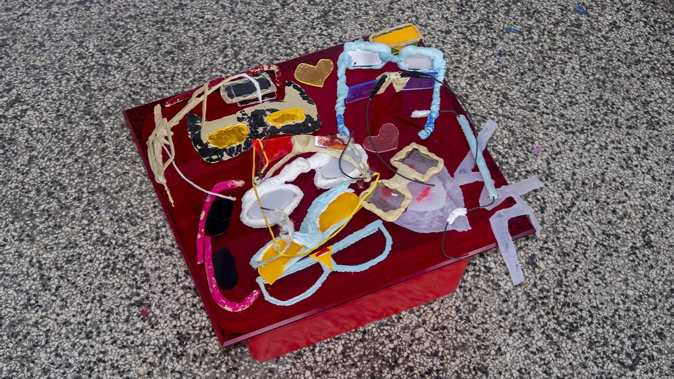 Collection of handmade artistic sunglasses and paper frames on a red glass table surface.