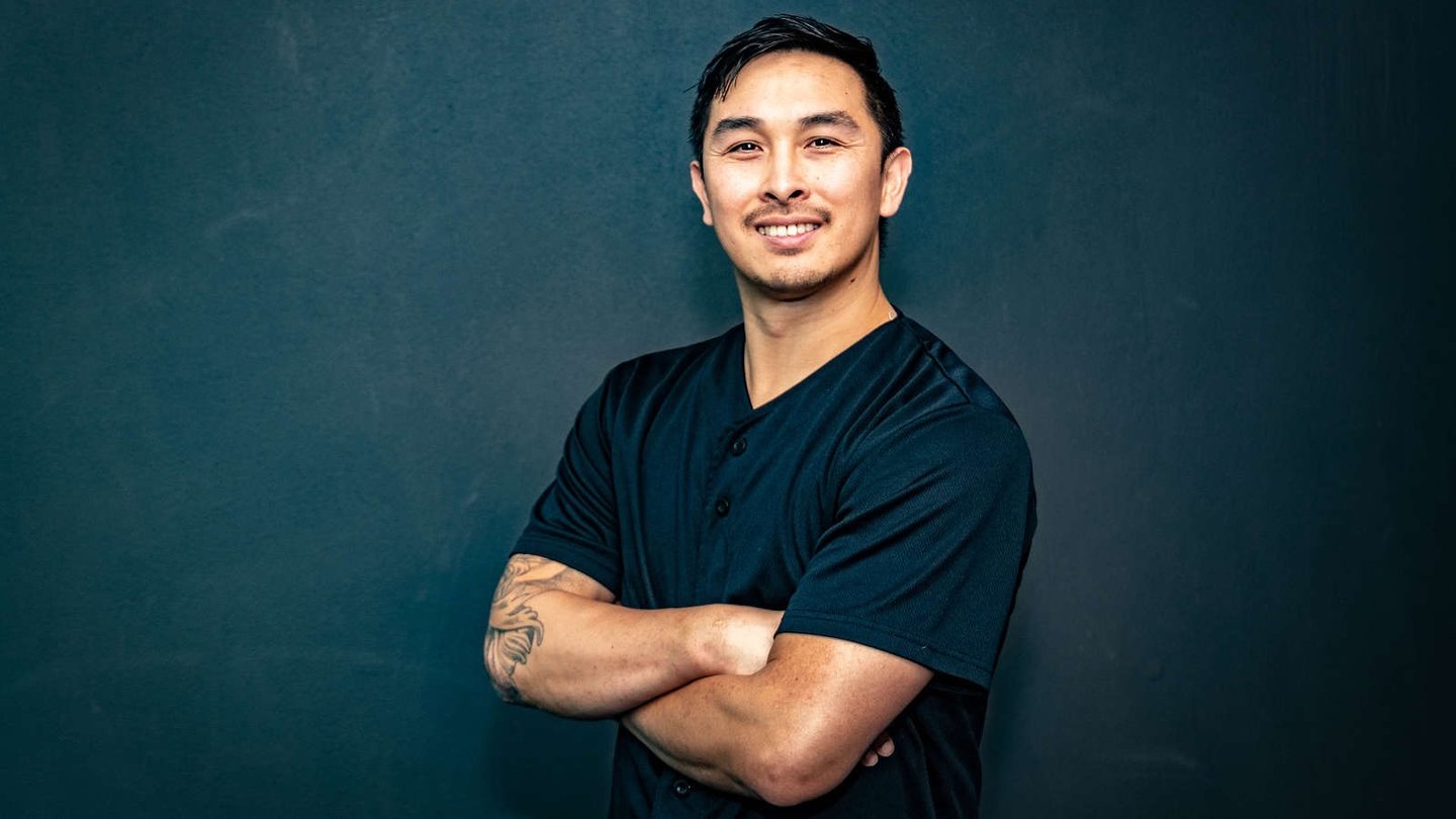 Smiling man with arm tattoos posing in a black shirt against a dark studio background.