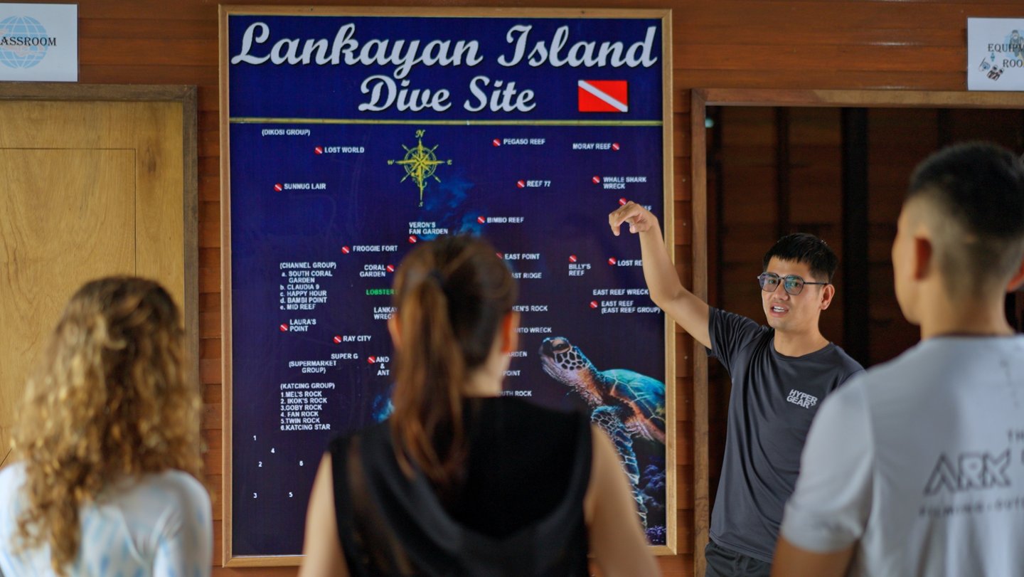 Dive guide giving a dive briefing to three divers at a site map at Lankayan Island