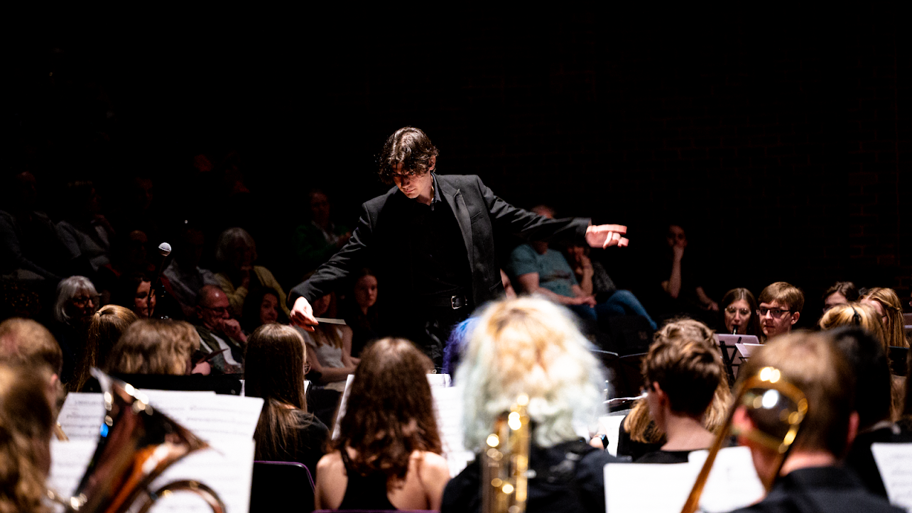 Conductor in black, with arms spread, amid heads of orchestra-members.
