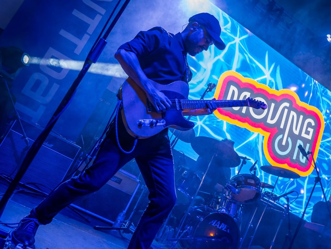 Foto real del guitarrista de Moving On tocando en un evento y el logo proyectado en pantalla