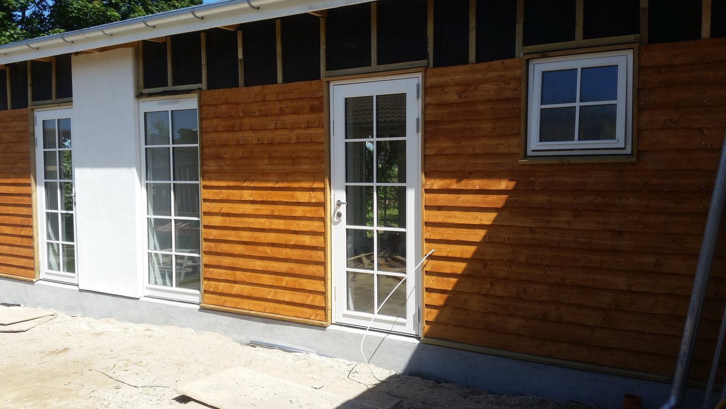Moderne havebygning under opførelse med vandret træbeklædning og hvide glasdøre.
