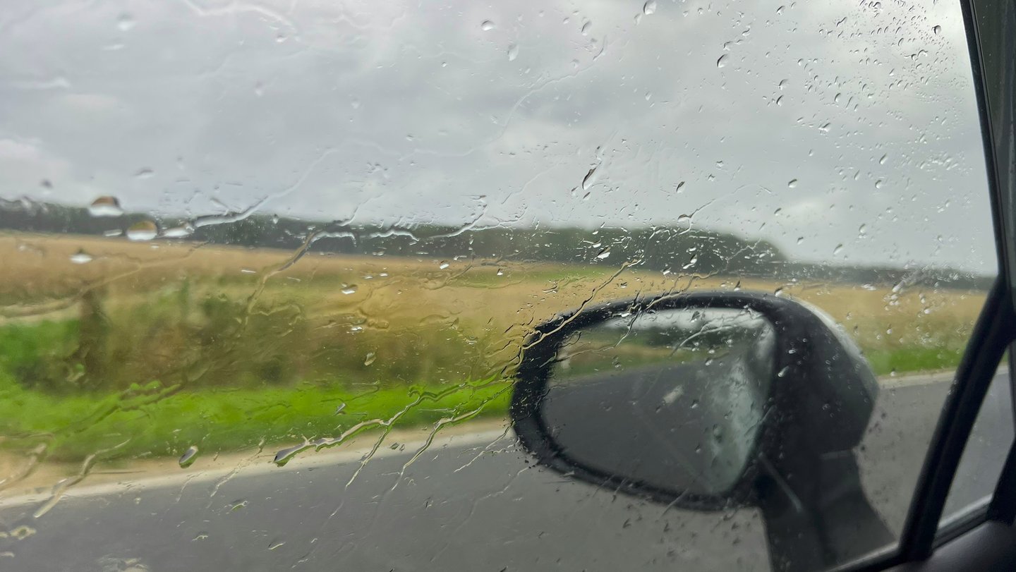 Blik vanuit auto door beslagen zijruit, met regendruppels en bos op achtergrond.