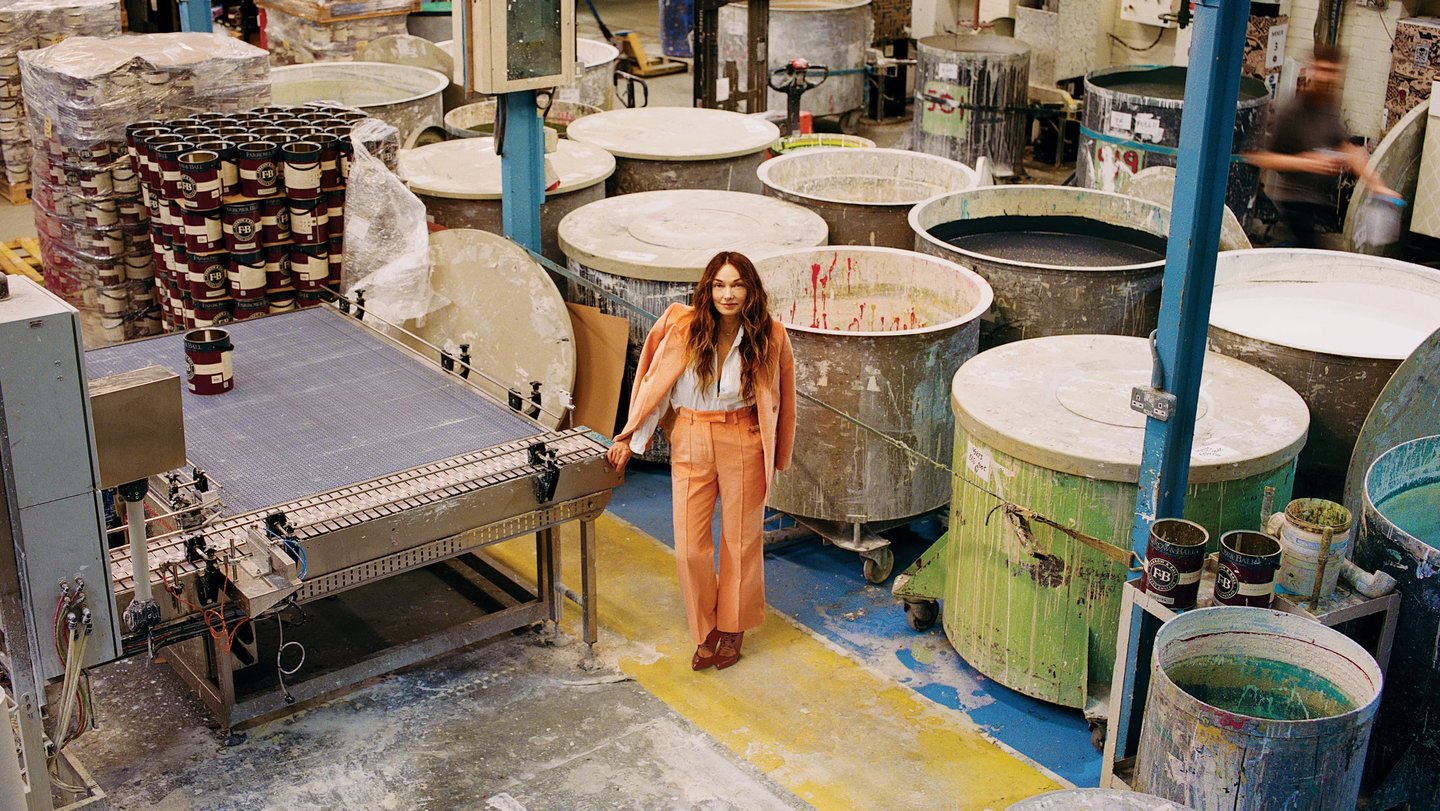 A woman in a coral suit standing in a professional paint factory with industrial mixing vats and conveyors.