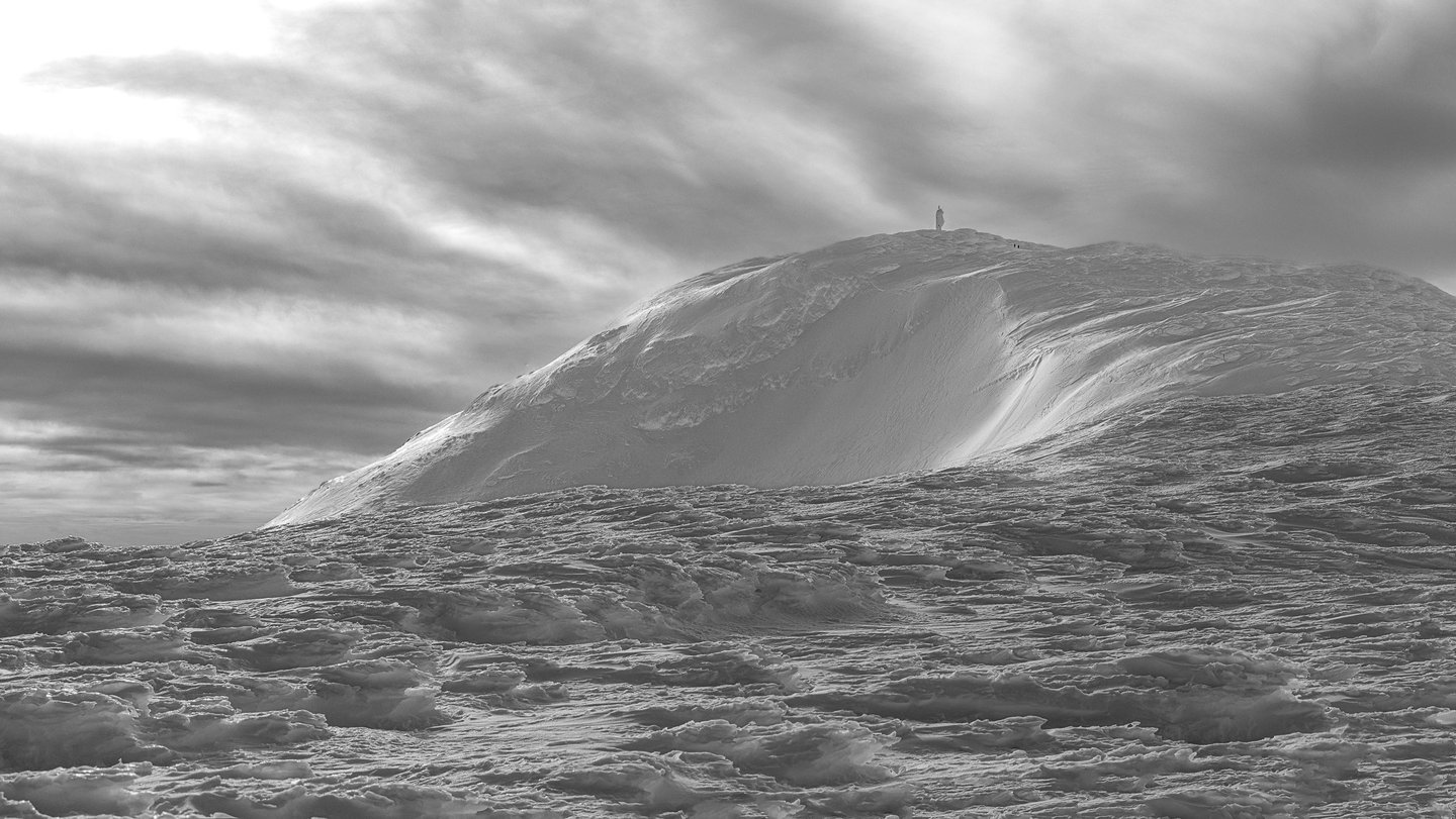 Black and white landscape of a lone hiker standing on a snowy mountain peak under a dramatic sky.