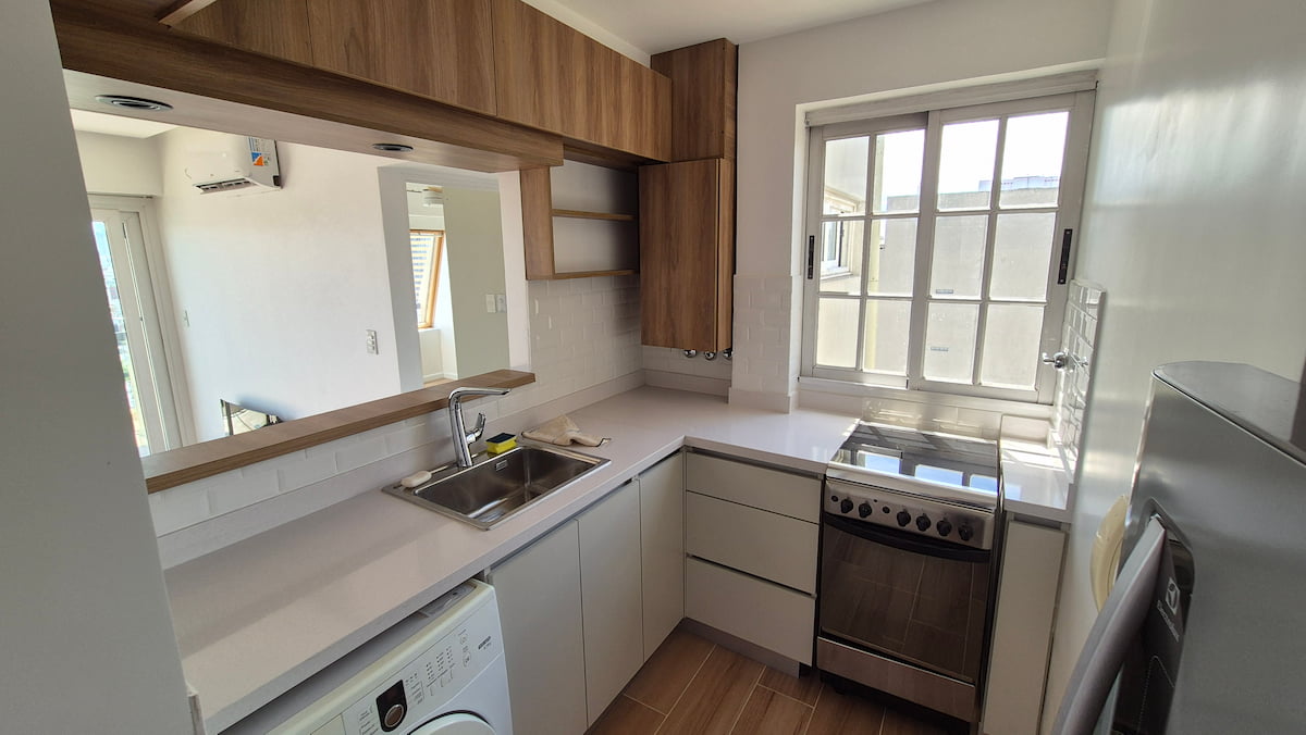 Modern small apartment kitchen with white countertops, wooden cabinets, and stainless steel appliances.