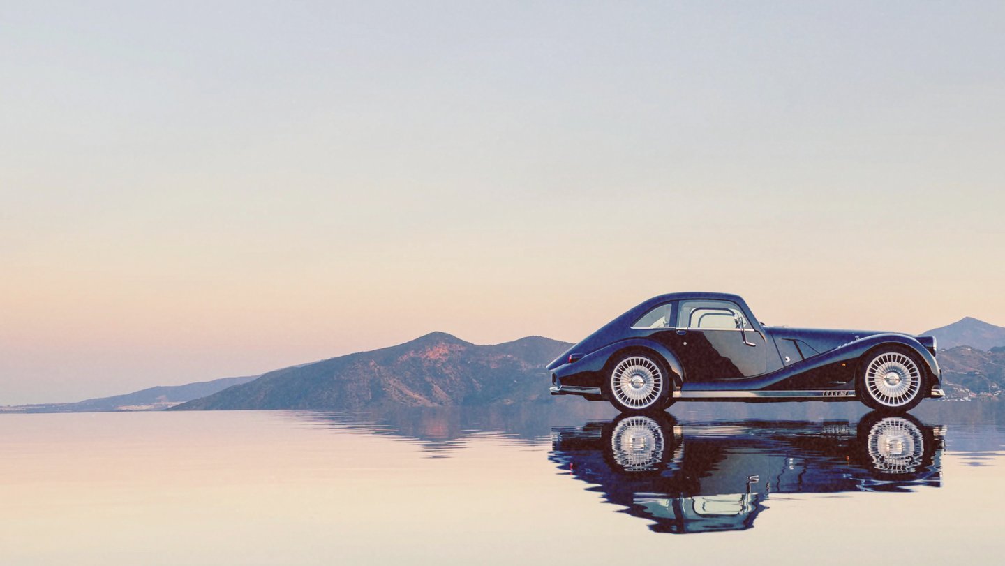 A vintage black luxury car reflecting on a calm water surface with mountains and a sunset sky in the background.
