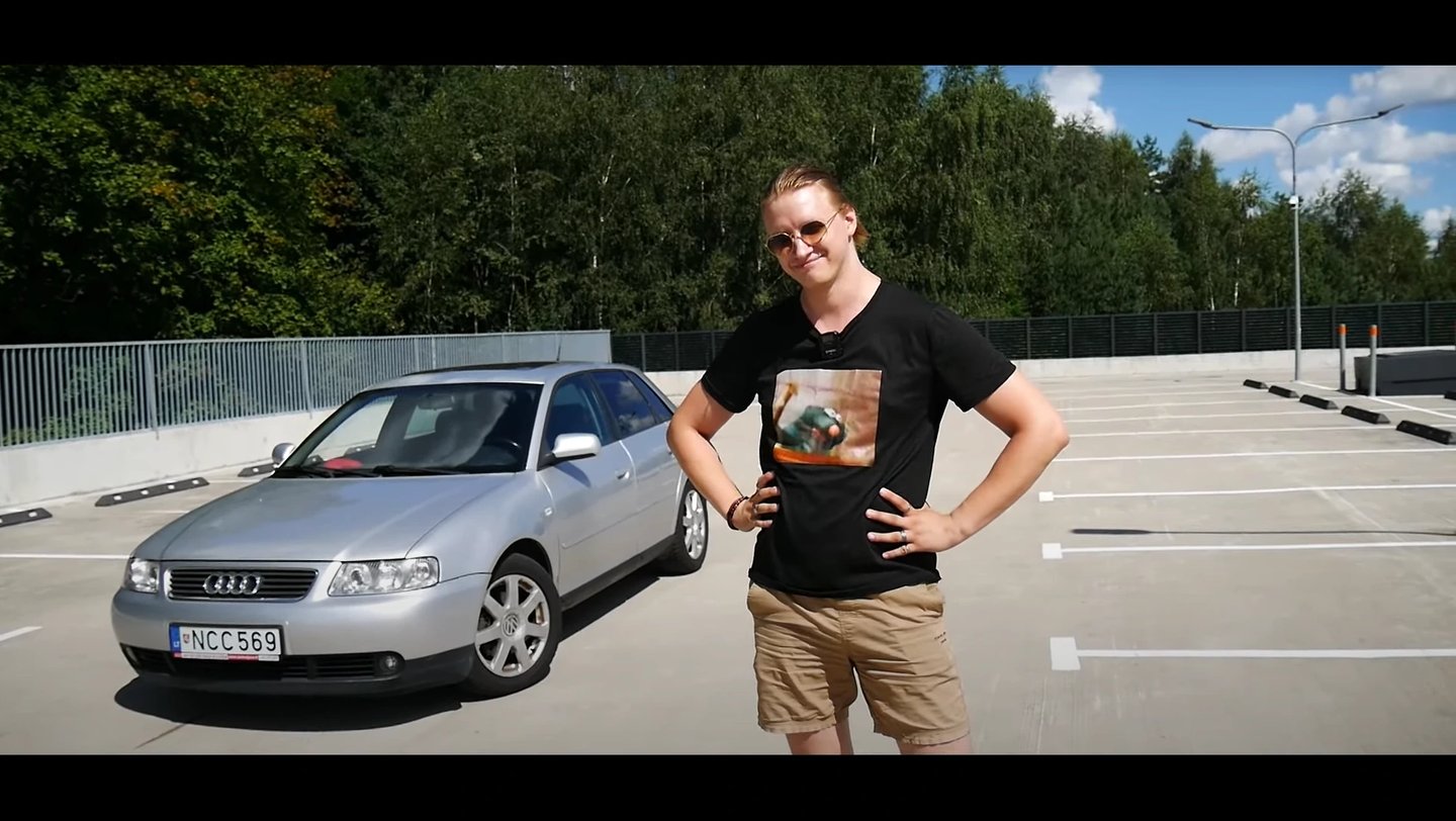 Youtuberis su juodais marškinėliais šalia sidabrinio Audi A3 saulėtoje aikštelėje.