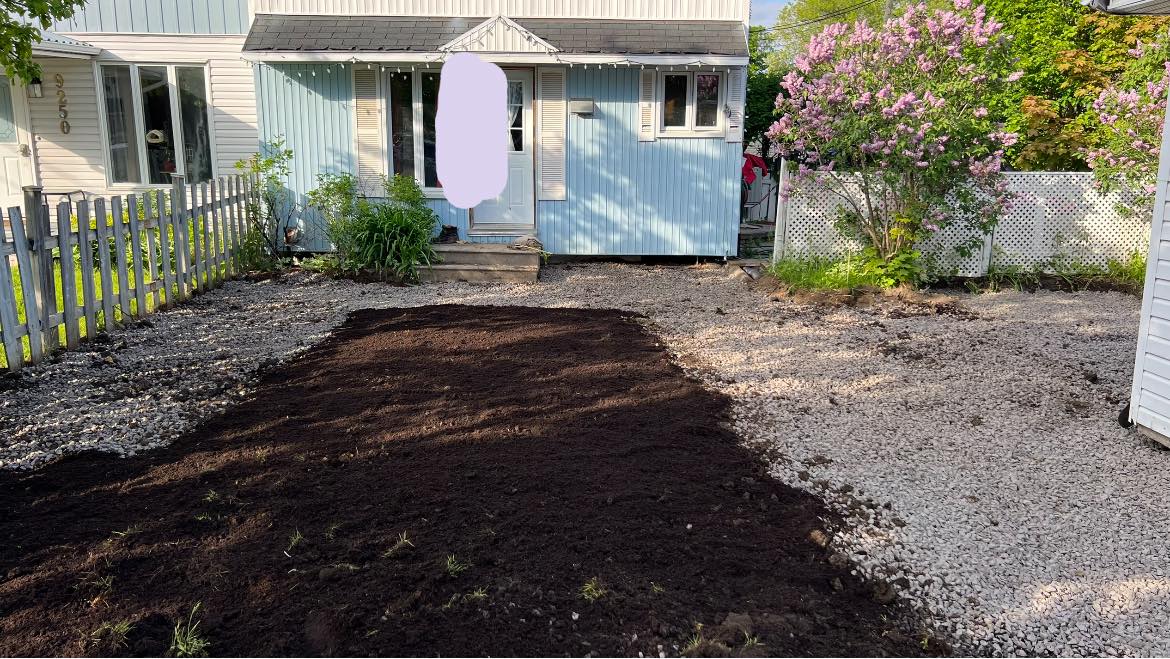 Fresh dark garden soil spread for landscaping in a gravel front yard of a blue house with pink flowers.