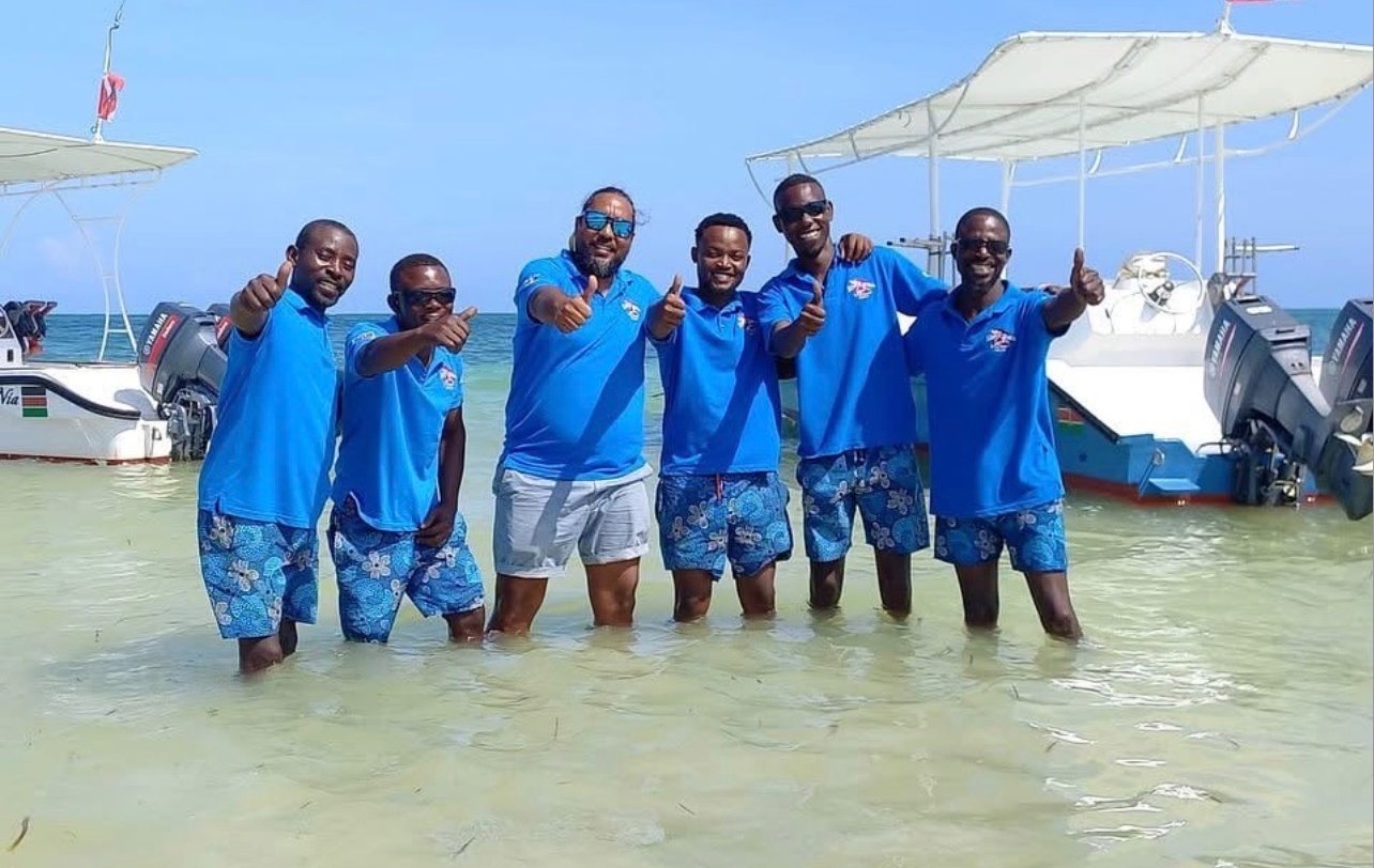 A group of diving instructors in blue shirts giving thumbs up in tropical ocean water near boats.
