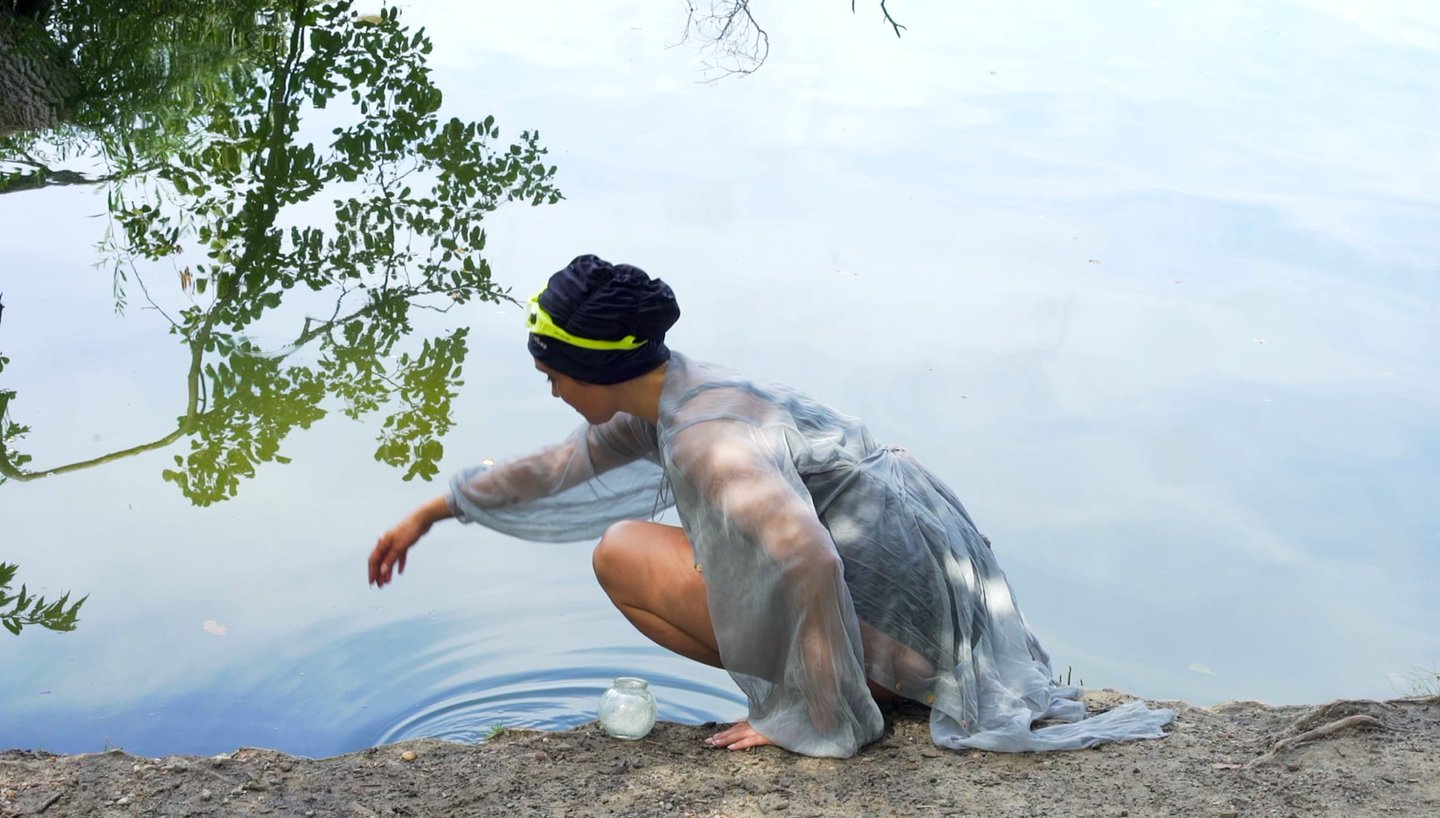 ilm still from "Fear" by Tina Bikic of a performer in a sheer garment crouching by the lake, reaching to the water.