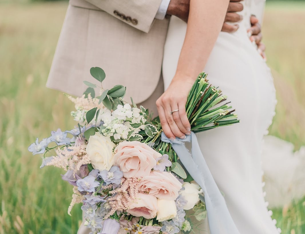a bride and groom standing in a field, the bride is holding a pastel flower wedding bouquet