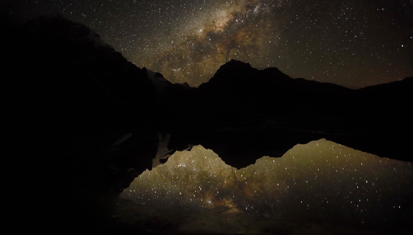 This image shows a serene night sky mirrored in calm water, likely featuring the Milky Way over a quiet mountain scene.